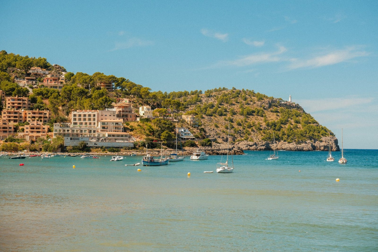 Boote liegen in einer Bucht auf Mallorca, im Hintergrund eine Küstenstadt auf einem Hügel sichtbar, aufgenommen von Alexis Presa.