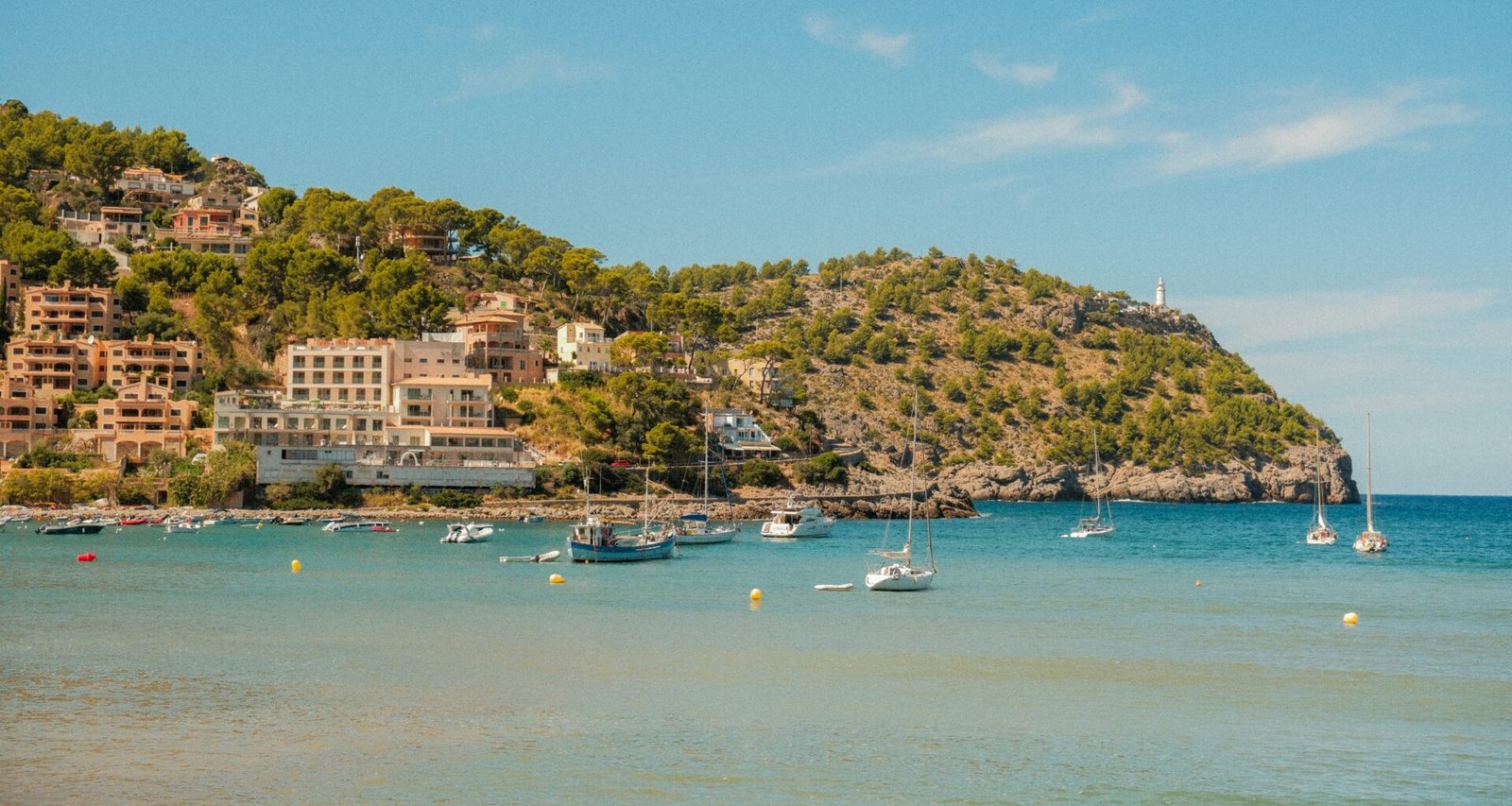 Boote liegen in einer Bucht auf Mallorca, im Hintergrund eine Küstenstadt auf einem Hügel sichtbar