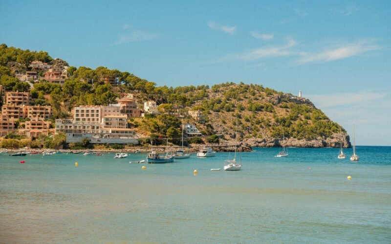 Boote liegen in einer Bucht auf Mallorca vor einer Küstenstadt auf einem Hügel, aufgenommen von Alexis Presa.
