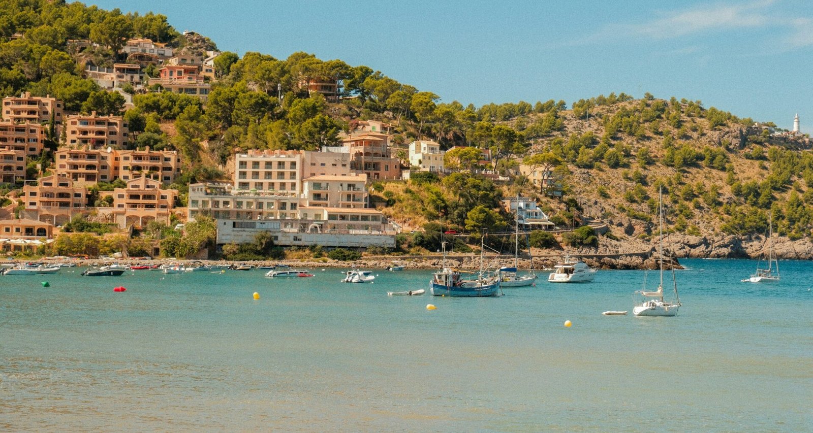 Boote liegen in einer Bucht auf Mallorca vor Häusern, die auf einem Hügel am Ufer gebaut sind, aufgenommen von Alexis Presa.
