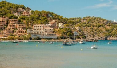 Boote liegen in einer Bucht auf Mallorca vor Häusern, die auf einem Hügel am Ufer gebaut sind, aufgenommen von Alexis Presa.