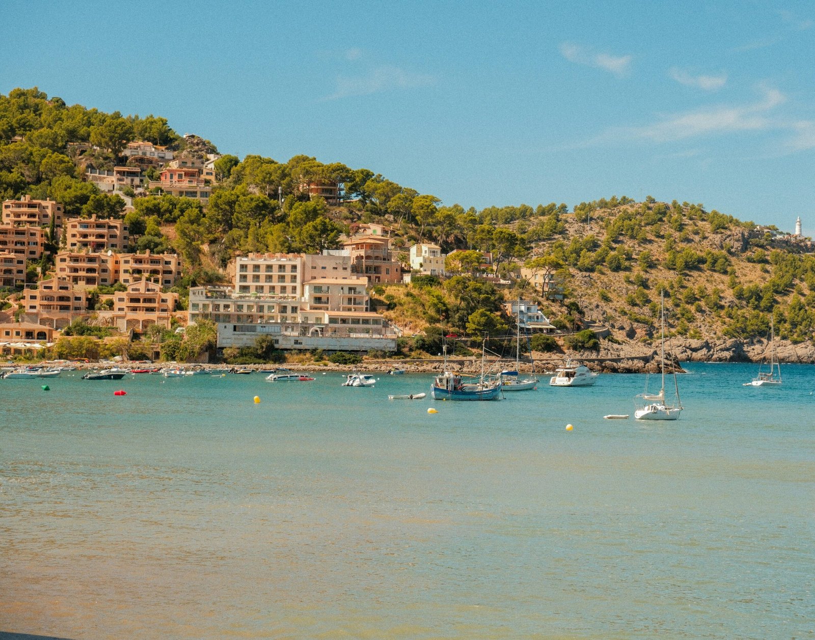 Boote liegen in einer Bucht auf Mallorca vor Häusern, die auf einem Hügel am Ufer gebaut sind, aufgenommen von Alexis Presa.