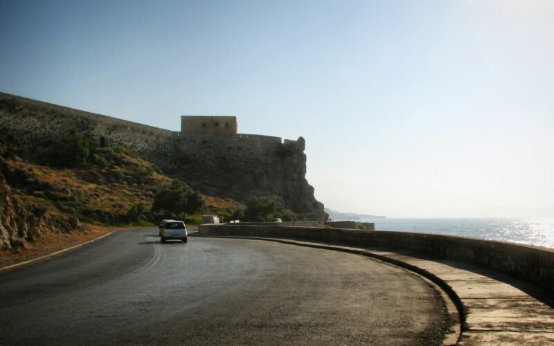 Ein Auto fährt auf Mallorca auf einer Küstenstraße in Richtung einer historischen Festung, aufgenommen von Sergei Gussev.