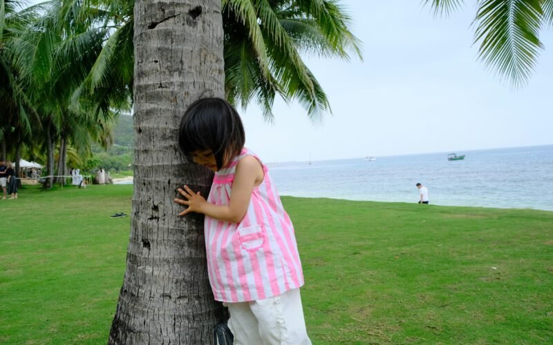 Kleines Mädchen versteckt sich hinter einer Palme auf Mallorca, umgeben von grünen Blättern und natürlicher Umgebung.