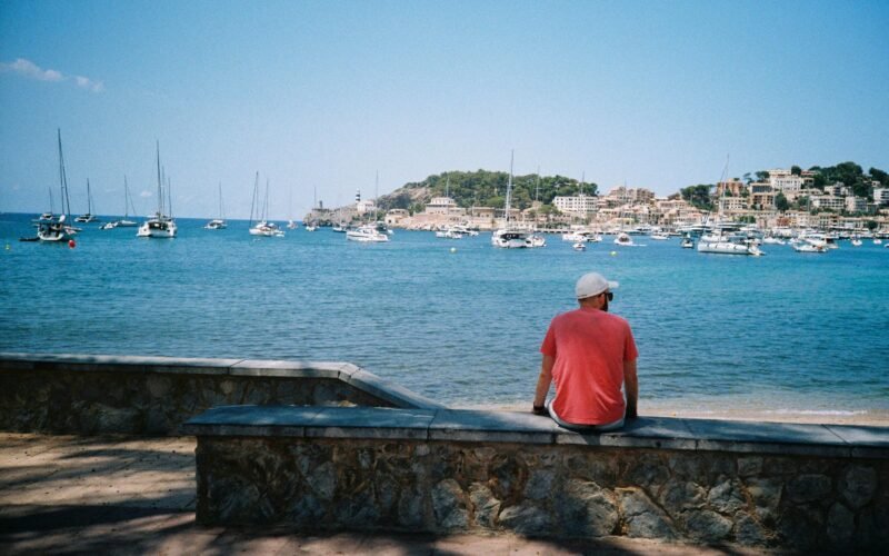 Mann sitzt auf Mallorca auf einer Mauer und blickt auf das Wasser, umgeben von natürlicher Kulisse