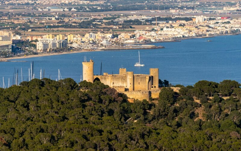Eine Burg auf einem Hügel auf Mallorca mit Blick auf ein ruhiges Gewässer, aufgenommen von David Vives.