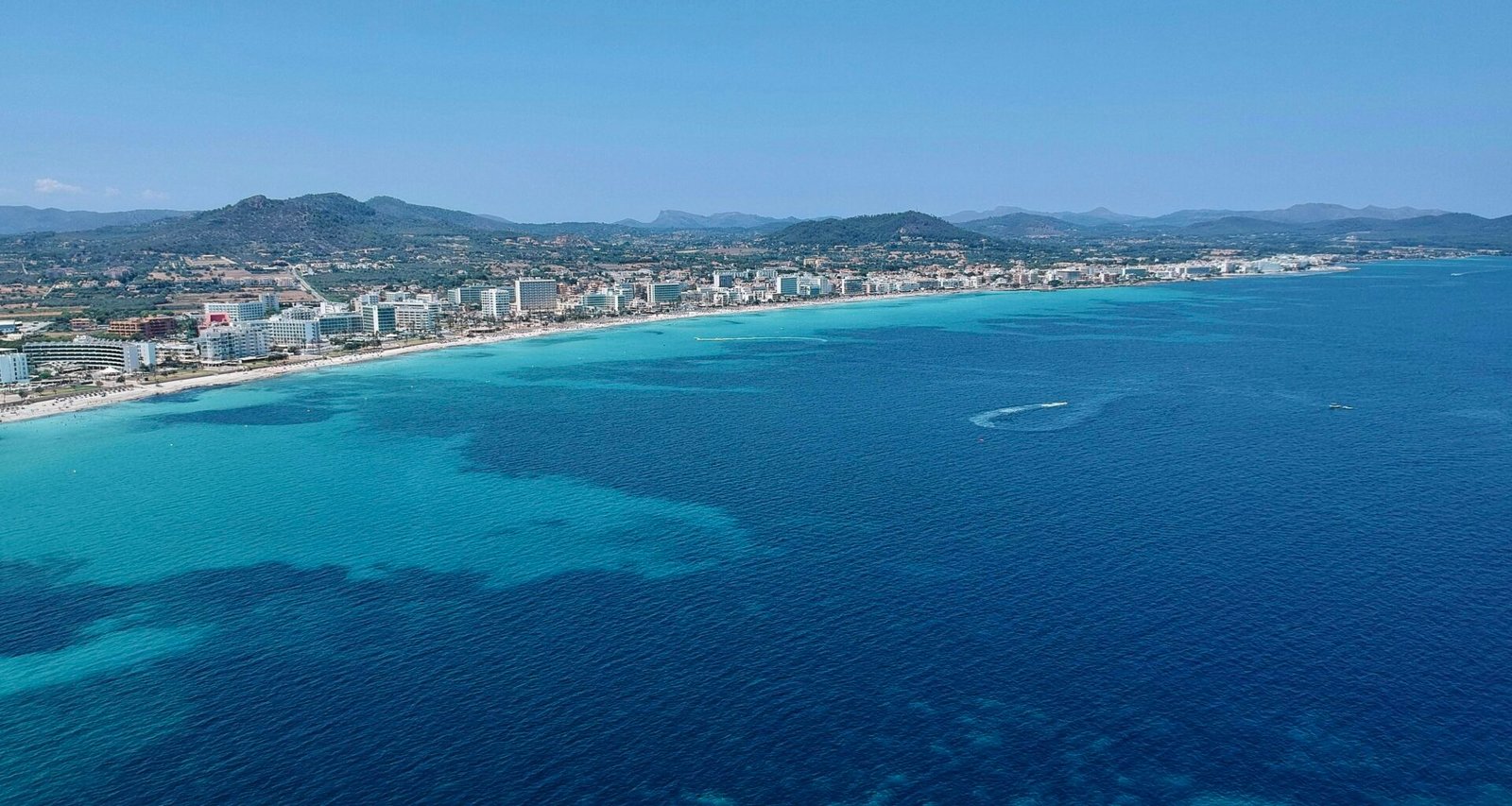 Stadt auf dem Wasser auf Mallorca mit Gebäuden am Ufer und einem ruhigen Wasser spiegelnd, aufgenommen von Leon Rohrwild.