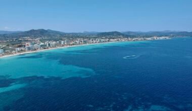 Stadt auf dem Wasser auf Mallorca mit Gebäuden am Ufer und einem ruhigen Wasser spiegelnd, aufgenommen von Leon Rohrwild.