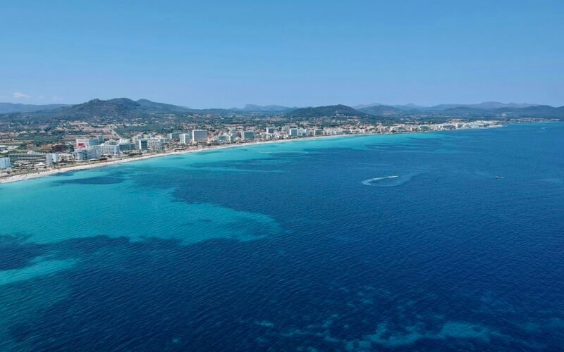 Stadt auf dem Wasser auf Mallorca mit Gebäuden am Ufer und einem ruhigen Wasser spiegelnd, aufgenommen von Leon Rohrwild.