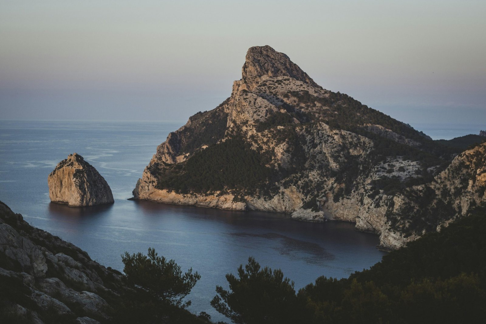 Felsige Küste mit Inseln und ruhigem Meer bei Dämmerung auf Mallorca, Sonnenuntergang am Horizont
