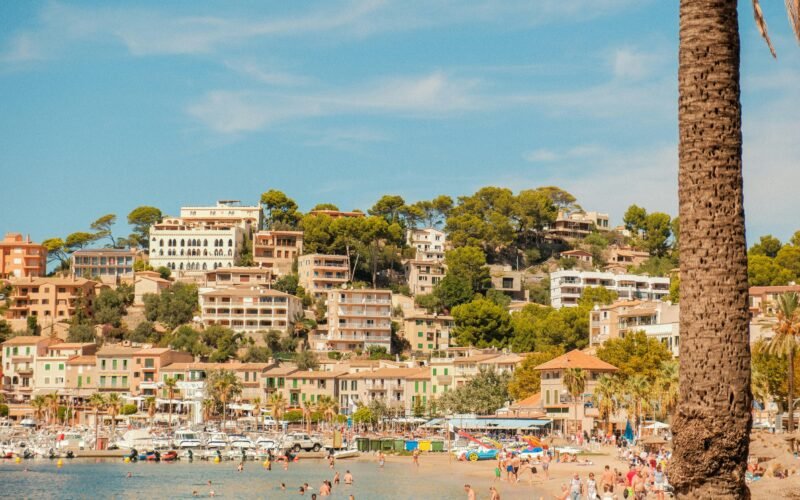 Küstendorf auf Mallorca mit Gebäuden auf einem Hügel, Blick auf den Strand im Vordergrund, aufgenommen von Alexis Presa