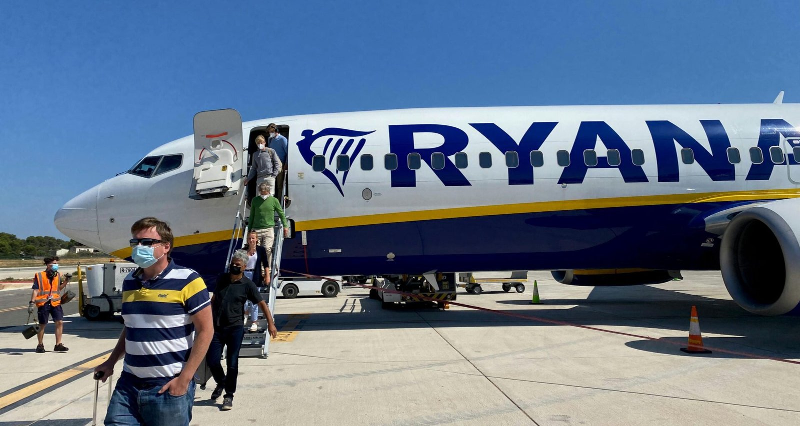 Mann in gelbem Hemd und grauen Shorts steht neben einem weißen und blauen Flugzeug auf Mallorca bei Tageslicht.