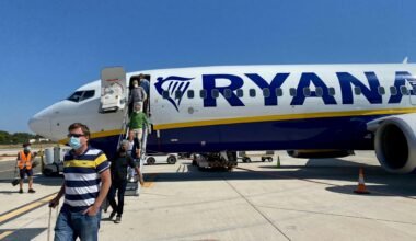 Mann in gelbem Hemd und grauen Shorts steht neben einem weißen und blauen Flugzeug auf Mallorca bei Tageslicht.