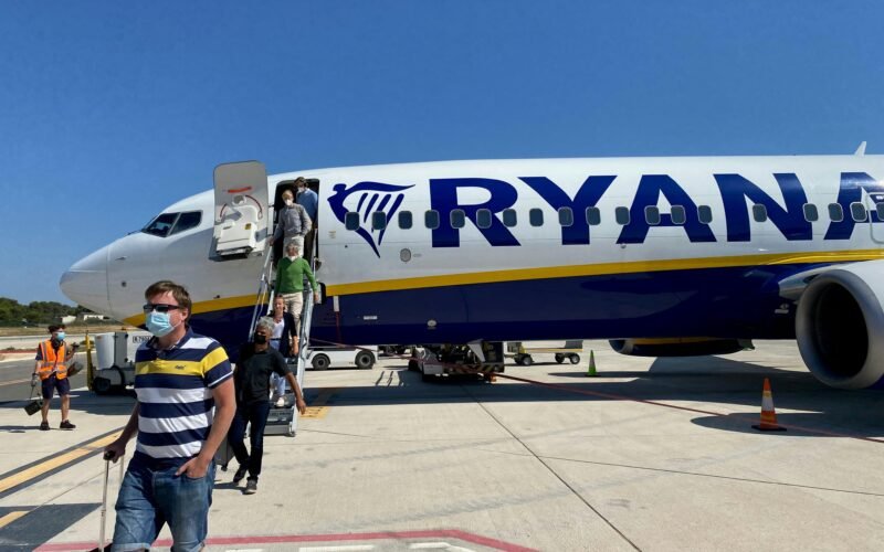 Mann in gelbem Hemd und grauen Shorts steht neben einem weißen und blauen Flugzeug auf Mallorca bei Tageslicht.
