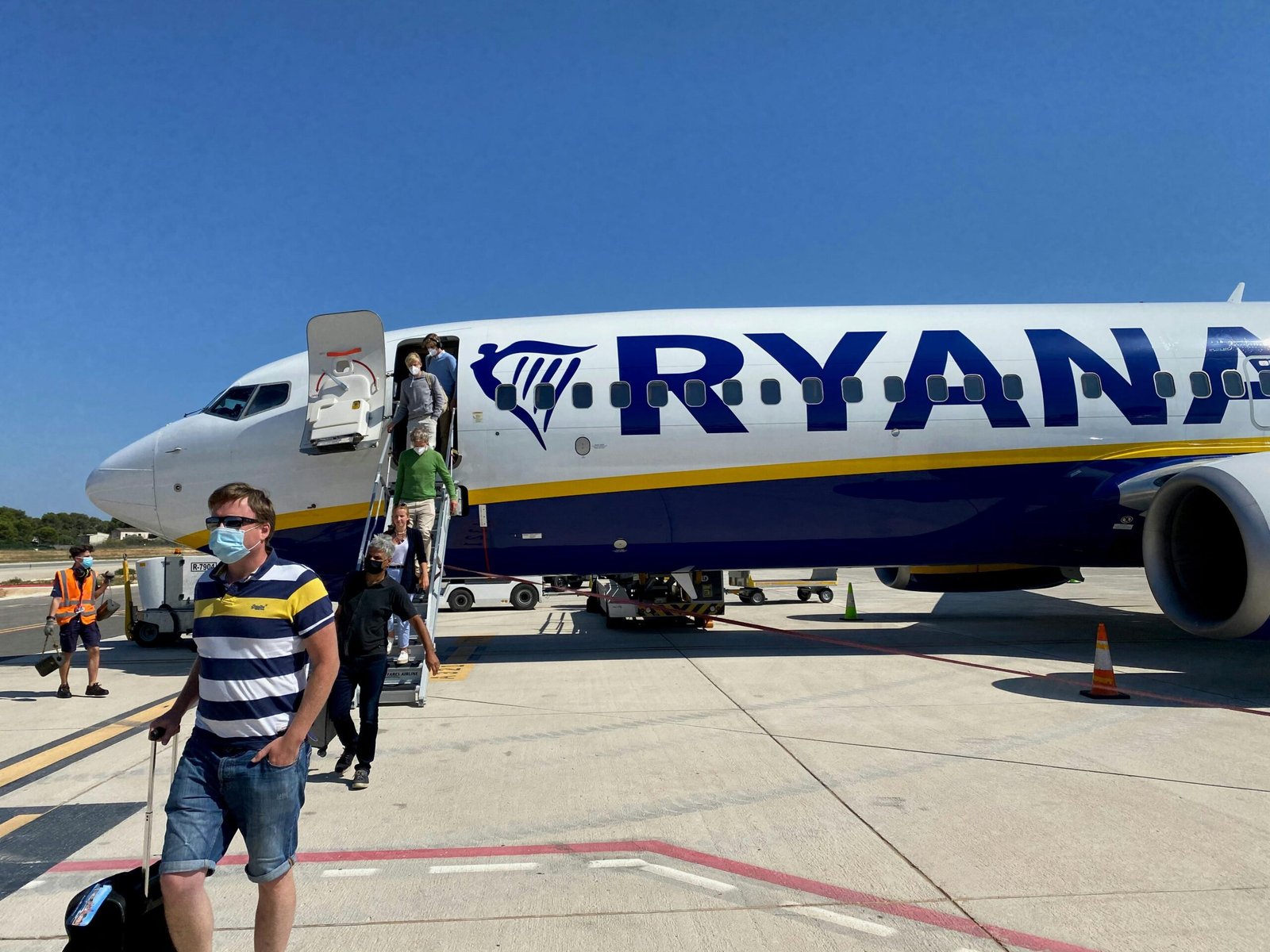 Mann in gelbem Hemd und grauen Shorts steht neben einem weißen und blauen Flugzeug auf Mallorca bei Tageslicht.