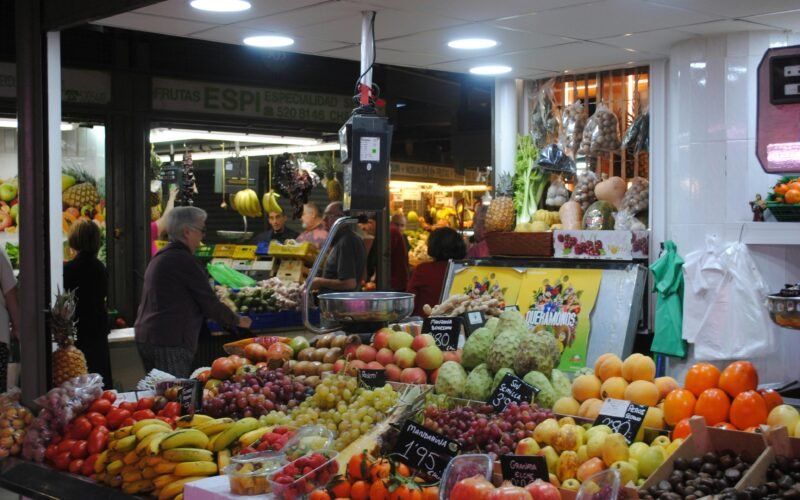 Mann in grauer Jacke steht neben Obststand auf Mallorca, umgeben von frischen Früchten