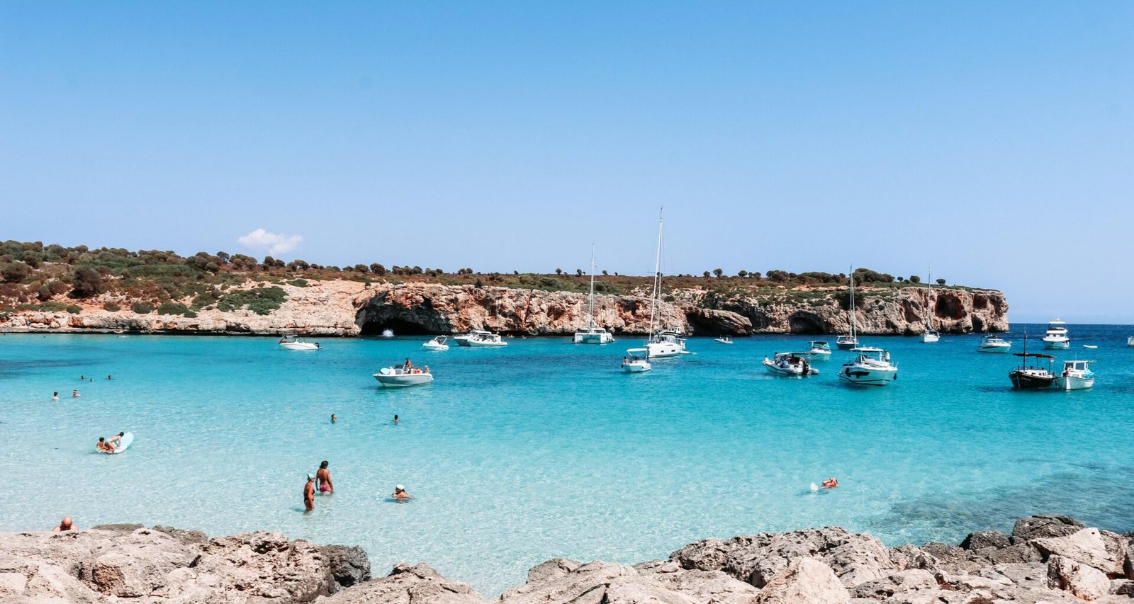Menschen schwimmen tagsüber am Strand auf Mallorca, das klare Wasser und der Sand sind sichtbar, aufgenommen von Austin Farrington.