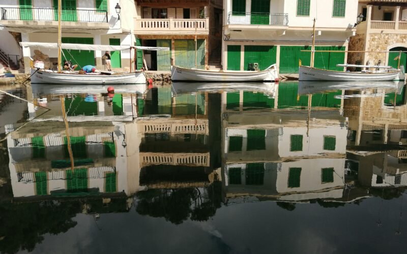 Weiße Boote liegen neben Gebäuden auf Mallorca auf dem Wasser, aufgenommen von Jacques Leideck.