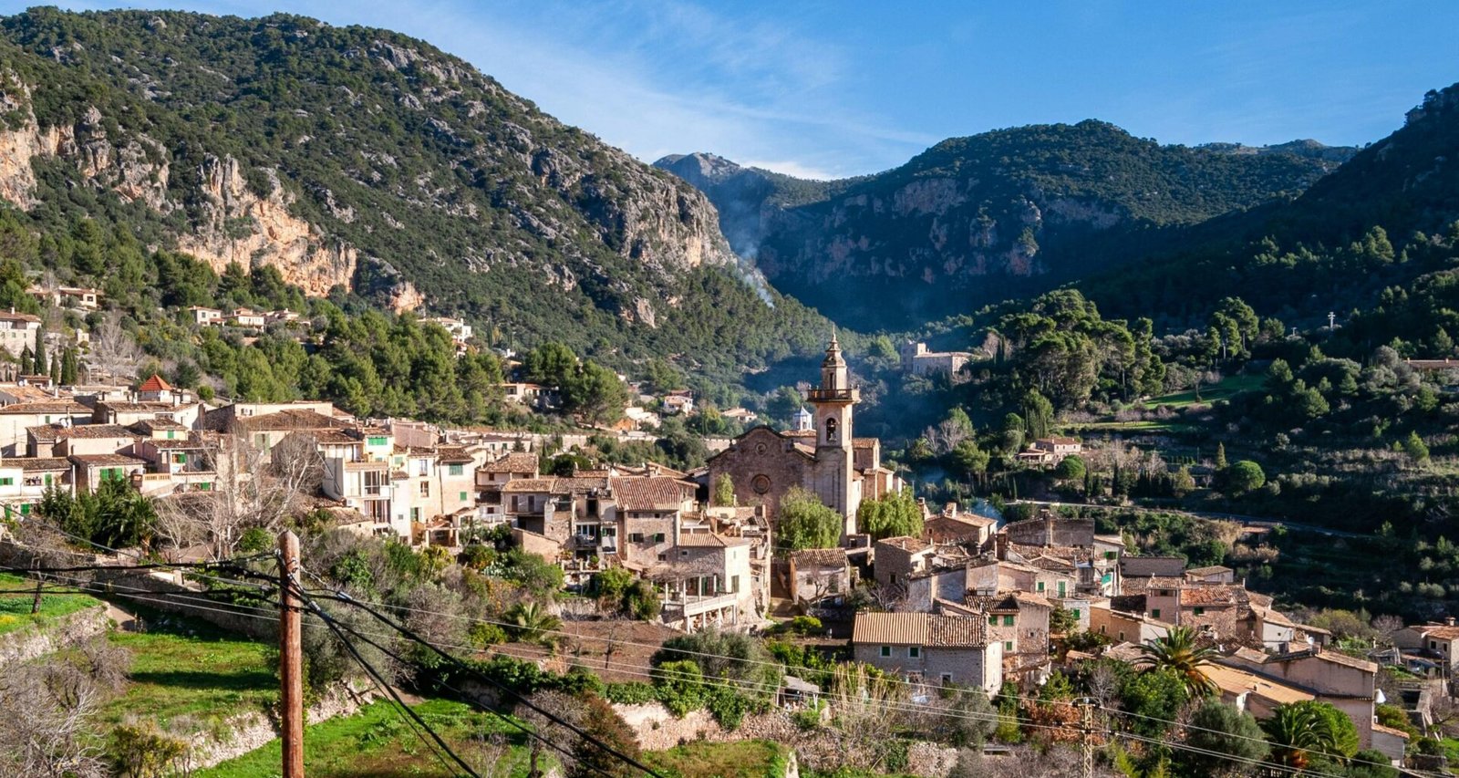 Weiße und braune Betonhäuser auf Mallorca vor grünen Bergen unter blauem Himmel während des Tages, fotografiert von David Vives.