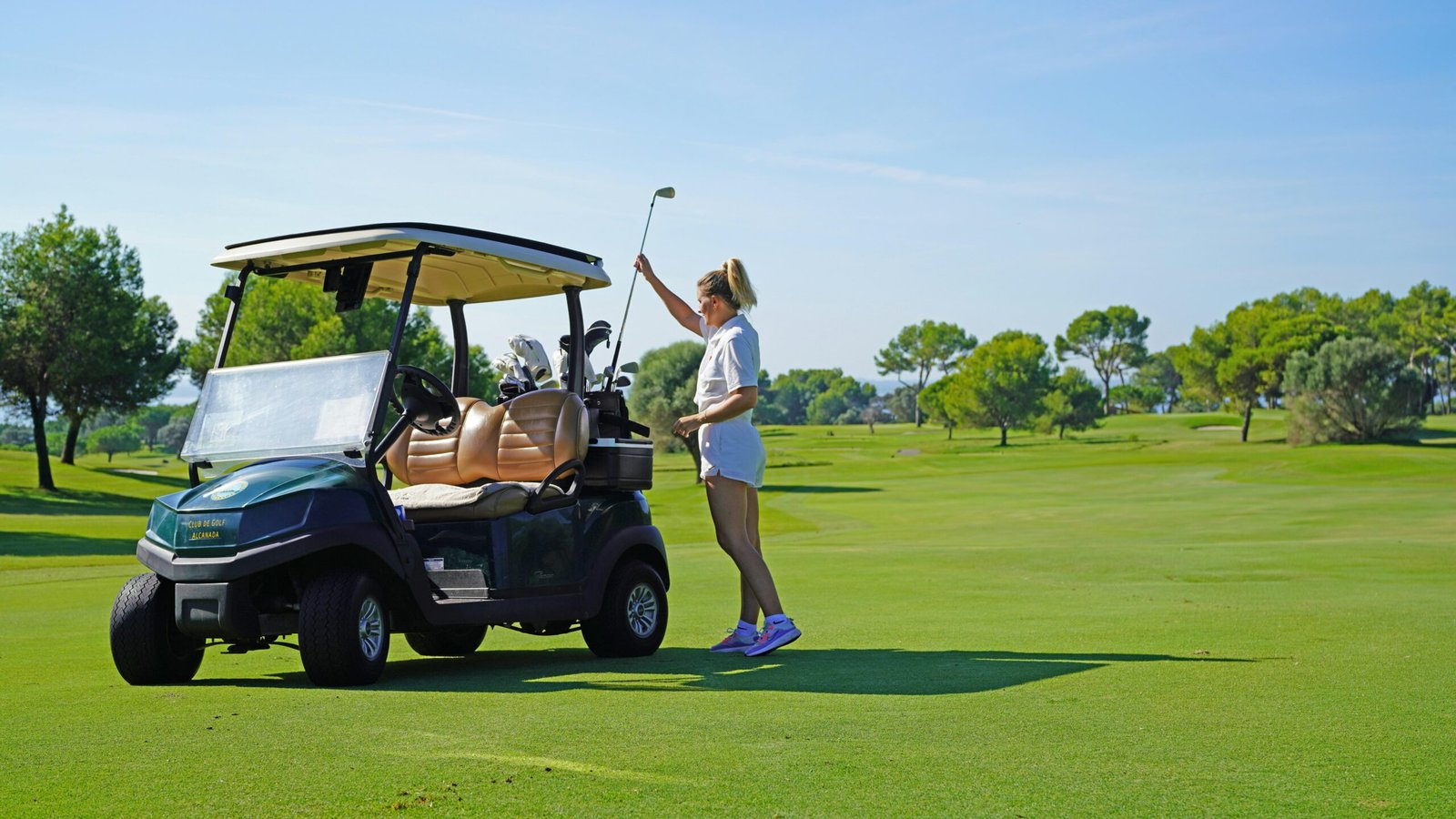 Frau steht neben einem Golfwagen auf einem Golfplatz auf Mallorca, umgeben von grünen Fairways und Bäumen.