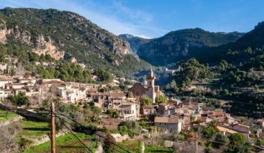 Weiße und braune Betonhäuser auf Mallorca vor grünen Bergen unter blauem Himmel während des Tages, fotografiert von David Vives.